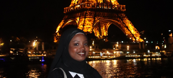 Rayhana Barry standing in front of the Effiel Tower at night while it is illumniated by yellow lights and the olympic rings..