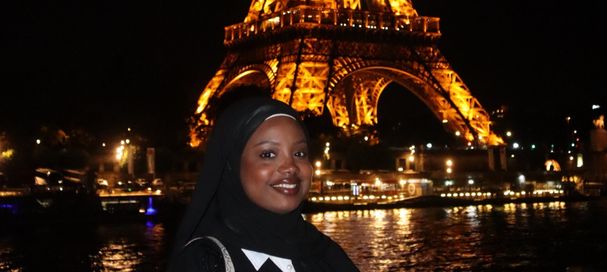 Rayhana Barry standing in front of the Effiel Tower at night while it is illumniated by yellow lights and the olympic rings..