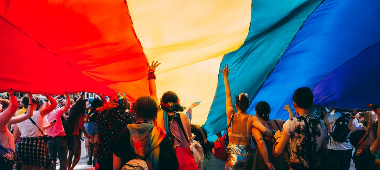 Individuals stand under a large LGBTQ+ flag during a pride parade. 