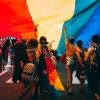 Individuals stand under a large LGBTQ+ flag during a pride parade. 
